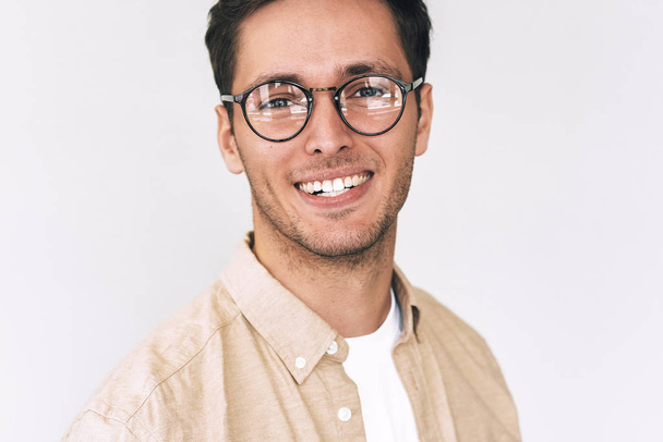 Close up portrait of cheerful young man, looks directly at the camera, wears round spectacles, isolated over white wall. Smart bristle student male smiling and wearing casual outfit. People emotions - Fotografie, Obrázek