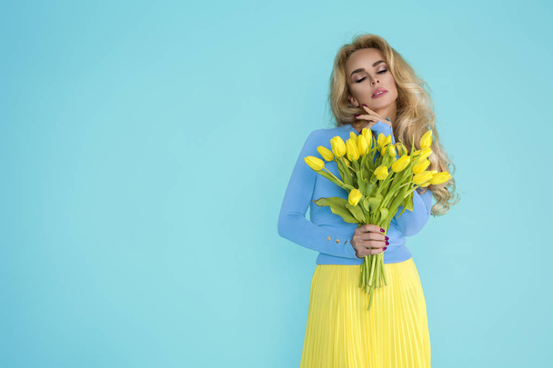 Весняна жінка. Краса Літня модель дівчини з барвистим одягом, що тримає букет весняних квітів. Красива дама з жовтими тюльпанами на синьому фоні
. - Фото, зображення