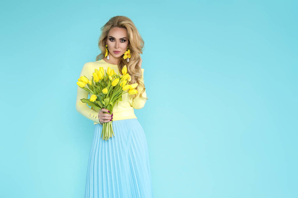 Spring Woman. Beauty Summer model girl with colorful clothes, holding a bouquet of spring flowers. Beautiful lady with yellow tulips on the blue background. - Photo, Image