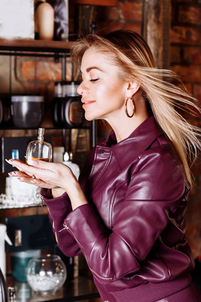 Beauty, aroma, people and body care concept. Young attractive woman with blond flying hair wearing violet leather jacket holding perfume in her hands - Фото, зображення