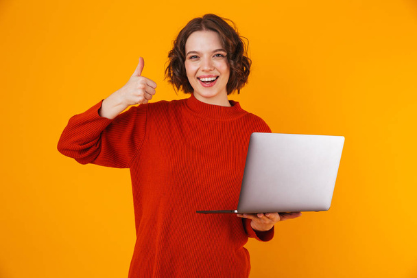 Excited emotional young pretty woman posing isolated over yellow wall background using laptop computer showing thumbs up. - Foto, Imagem