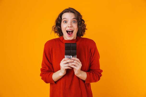 Emotional young pretty woman posing isolated over yellow wall background holding chocolate. - Фото, зображення