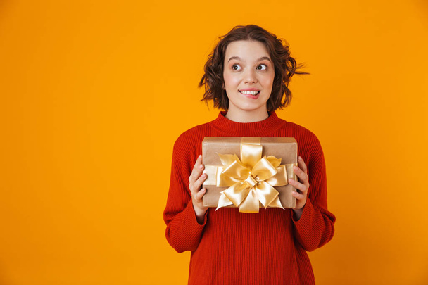 Emotional young pretty woman posing isolated over yellow wall background holding present gift. - Photo, Image