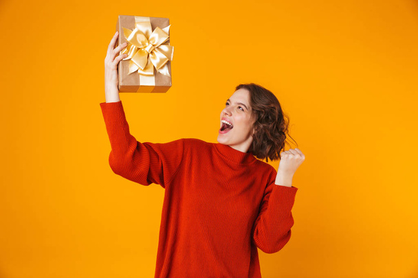 Emotional young pretty woman posing isolated over yellow wall background holding present gift. - Foto, afbeelding