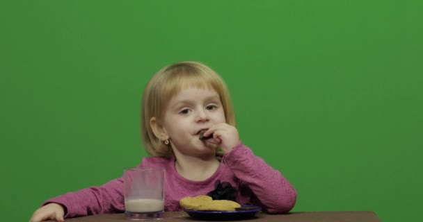 Dziewczyna siedzi przy stole i jedzenia czekolady i ciastek. Szczęśliwy trzy lat dziewczyna. Ładna dziewczyna uśmiechając się. Dość małe dziecko, 3-4-letniego Blondynka. Zrobić twarze. Zielony ekran wideo. Kluczowanie kolorem - Materiał filmowy, wideo