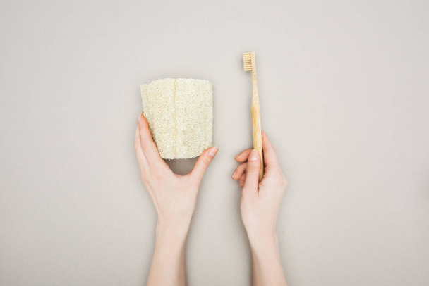 vista cortada de mulher segurando esponja orgânica e escova de dentes de bambu em fundo cinza
 - Foto, Imagem