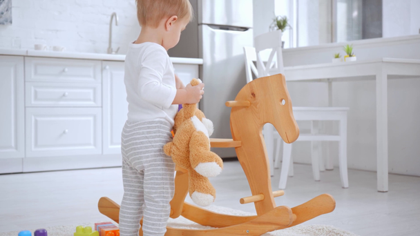 cute toddler boy holding teddy bear and playing with building blocks on rocking horse - Filmati, video