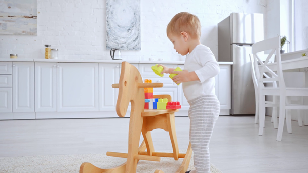 schattige peuter jongen spelen met plastic bouwstenen op schommelpaard in ruim appartement - Video
