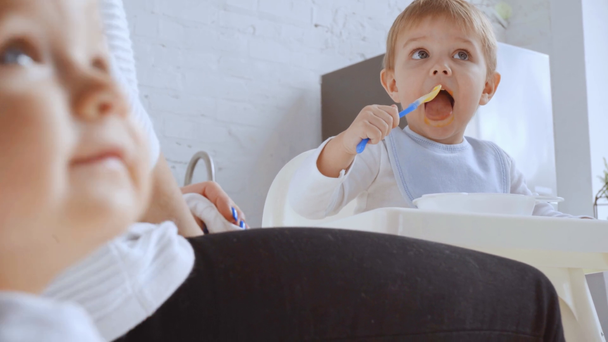 избирательный фокус ребенка указывая пальцем на брата, пока мальчик ест пюре в стульчике и мать вытирает рот салфеткой
 - Кадры, видео