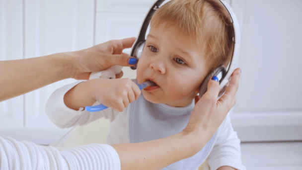 cute toddler boy sitting in highchair, eating puree and listening music in headphones - Footage, Video