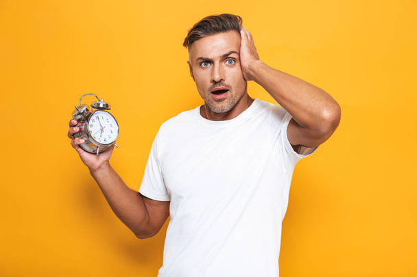 Confused man posing isolated over yellow wall background holding alarm clock. - Fotoğraf, Görsel