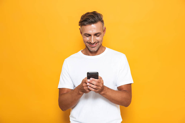Happy man posing isolated over yellow wall background using mobile phone. - 写真・画像