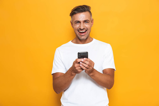 Happy man posing isolated over yellow wall background using mobile phone. - Photo, image