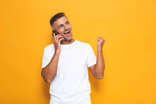 Emotional man posing isolated over yellow wall background talking by mobile phone. - 写真・画像