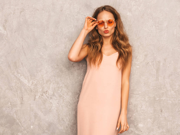 Portrait of young beautiful smiling hipster girl in trendy summer light pink dress. Sexy carefree woman posing near gray wall. Positive model having fun in round sunglasses - Fotó, kép
