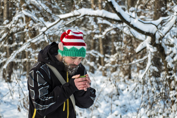 Homem barbudo em um chapéu de elfo na floresta aquece as mãos com uma caneca de chá quente
 - Foto, Imagem