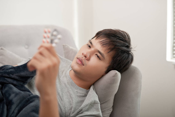 Un joven está enfermo de gripe, yace en casa bajo una manta, toma una pastilla
 - Foto, Imagen