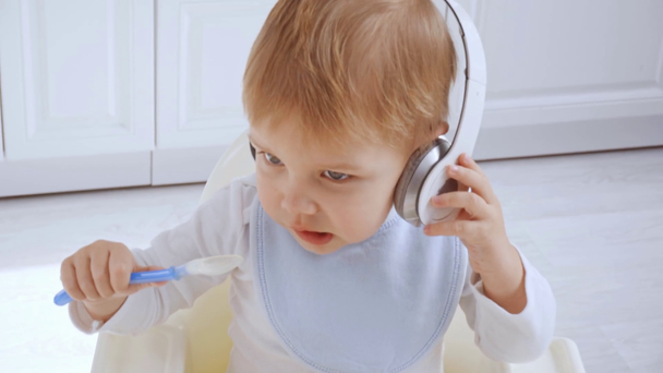 Niedlicher Kleinkind Junge sitzt im Hochstuhl, hält Plastiklöffel und hört Musik über Kopfhörer - Filmmaterial, Video