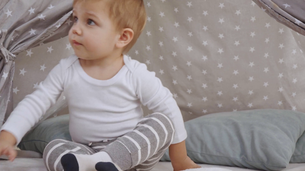 Niedlicher Kleinkind Junge sitzt in Baby-Perücke und spielt mit Buch, dann setzt Mutter Kopfhörer auf den Kopf und Baby liegt auf Teppich - Filmmaterial, Video
