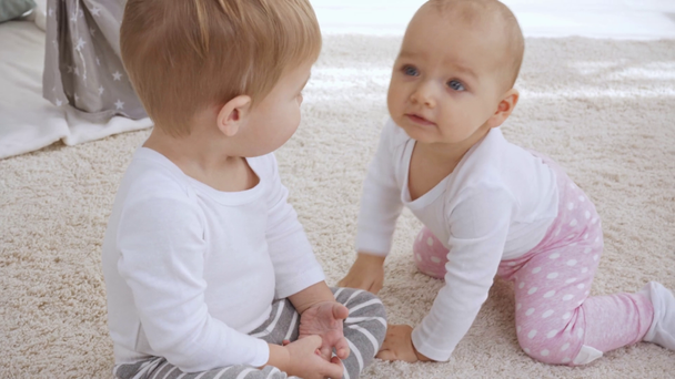 leuke jongen zittend op tapijt en wijzend met de vinger op de camera, terwijl de baby zus kruipen op handen en voeten en dan klap hands - Video