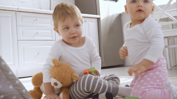 confused and smiling toddler boy sitting on carpet with teddy bear while baby sister touching his face - Imágenes, Vídeo