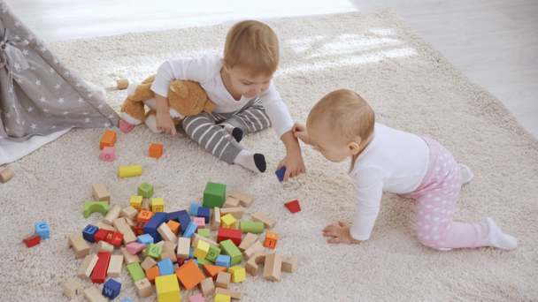 Малыш сидит на ковре с плюшевым мишкой и младшей сестрой и играет с деревянными блоками вместе
 - Кадры, видео