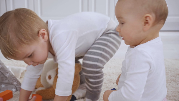 Kleinkind sitzt auf Teppich mit Teddybär, während die kleine Schwester mit Holzklötzen spielt, dann steht der Bruder auf und geht mit Spielzeug weg - Filmmaterial, Video