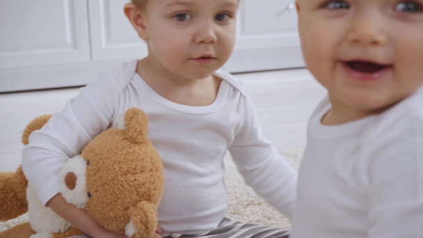 sad boy sitting on carpet with teddy bear while baby sister touching his face and taking wooden cubes from him - Footage, Video