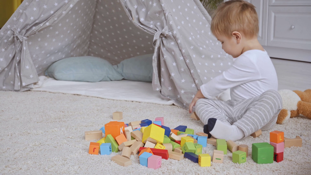 schattige peuter jongen bedrijf teddybeer, spelen met veelkleurige houten blokken op tapijt in de buurt van schommelpaard - Video