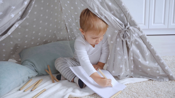 koncentrált kisgyermek fiú ül a szőnyegen a baba Wigwam és rajz színes ceruzák papíron - Felvétel, videó