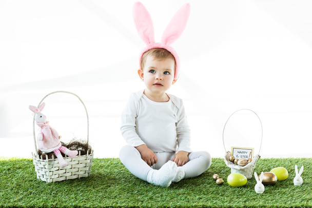 adorable enfant dans les oreilles de lapin bandeau assis près des paniers de paille avec des œufs colorés de Pâques et des lapins décoratifs isolés sur blanc
 - Photo, image