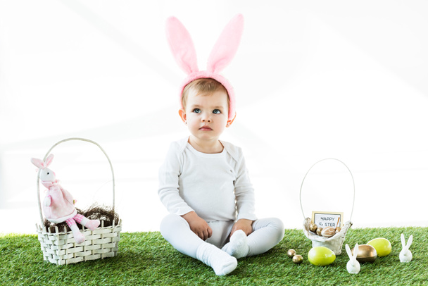 adorable bebé en orejas de conejo diadema sentado cerca de cestas de paja con huevos de Pascua coloridos y conejos decorativos aislados en blanco
 - Foto, Imagen
