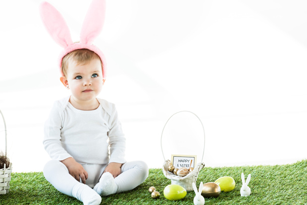 cute baby sitting near straw basket, Easter eggs and decorative rabbits isolated on white - Photo, Image