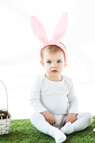lindo niño de ensueño en orejas de conejo diadema sentado en hierba verde cerca de la cesta de paja aislado en blanco
 - Foto, Imagen