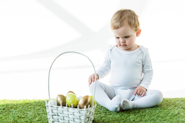 白い上に孤立した伝統的なエステルの卵とわらのバスケットの近くの緑の草の上に座っているかわいい赤ちゃん - 写真・画像