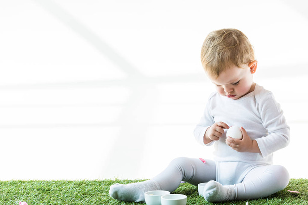 cute baby holding chicken egg while sitting on green grass near white bowls isolated on white - Foto, Bild