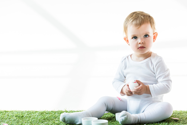 adorable baby sitting on green grass near white bowls and holding chicken egg isolated on white - Foto, imagen