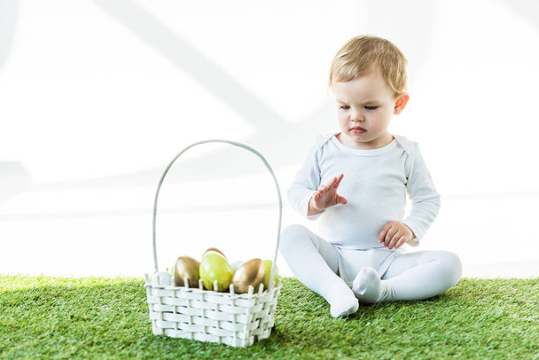adorable child sitting on green grass near straw basket with traditional Easter eggs isolated on white - Photo, Image