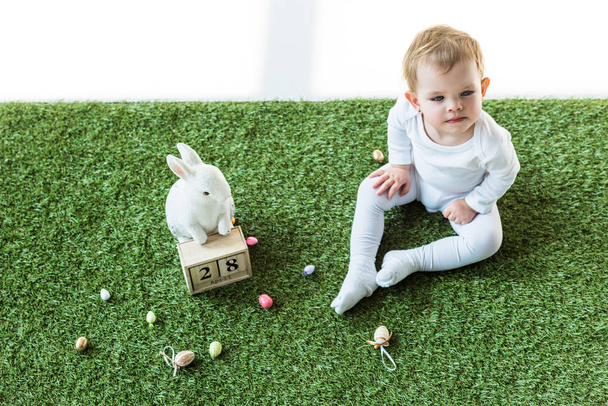 милый ребенок сидит на зеленой траве рядом с деревянным календарем с 28 апреля даты, декоративный кролик и красочные перепелиные яйца изолированы на белом
 - Фото, изображение