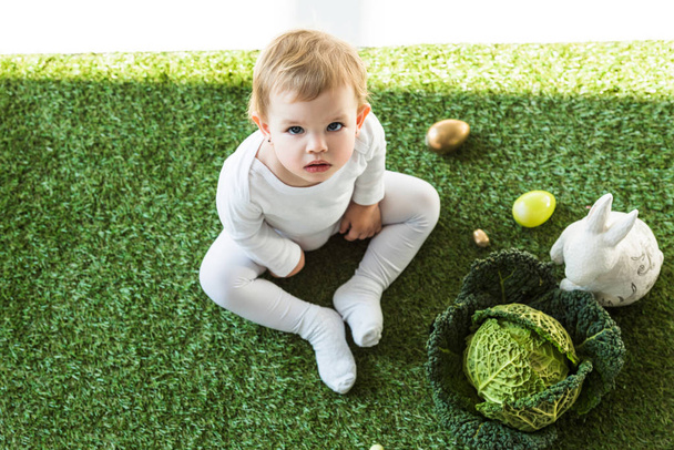overhead view of adorable child looking at camera while sitting near Easter eggs, decorative rabbit and savoy cabbage - Φωτογραφία, εικόνα