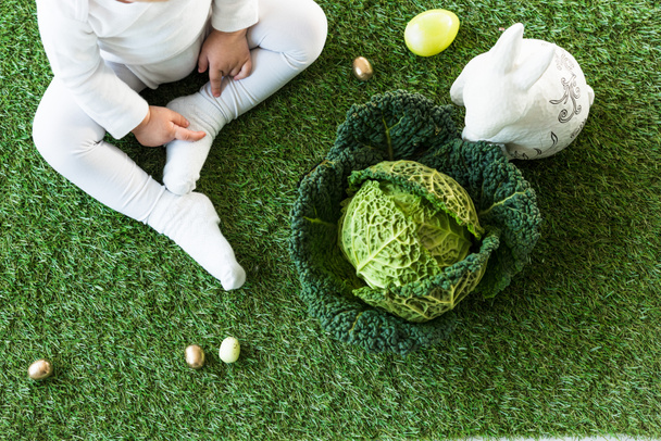vista parcial del niño sentado en la hierba verde cerca de los huevos de Pascua, conejo decorativo y col de savoy
 - Foto, Imagen