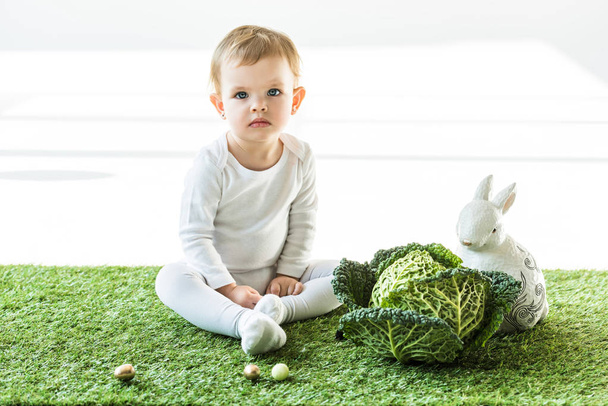 lindo bebé sentado cerca de col savoy, conejo decorativo y coloridos huevos de codorniz en blanco
 - Foto, Imagen