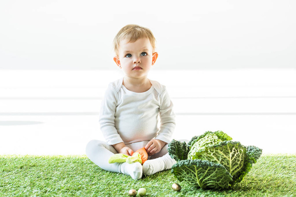 adorable kid sitting on green grass near golden quail eggs and savoy cabbage - Photo, Image
