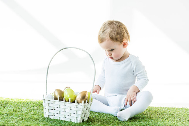 очаровательный ребенок сидит рядом с соломенной корзиной с блестящими пасхальными яйцами изолированы на белом
 - Фото, изображение