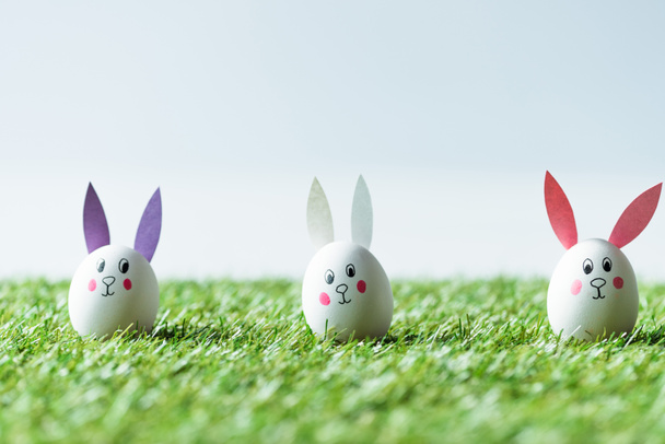 selective focus of Eastern chicken eggs with funny bunny faces and paper ears on green grass  - 写真・画像