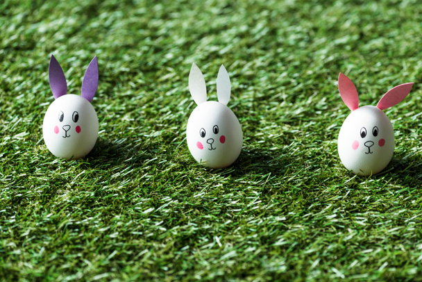 enfoque selectivo de huevos de pollo con caras de conejo dibujadas y orejas de papel en la superficie de hierba verde
 - Foto, Imagen