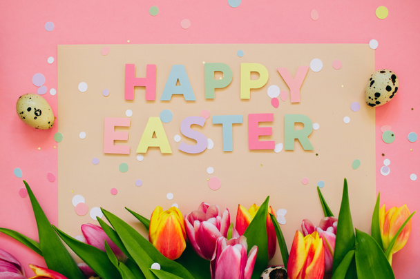 Easter concept. Happy easter paper cut inscription with pink and red tulips and quail eggs on pink background. Copy space, flat lay. - Photo, image