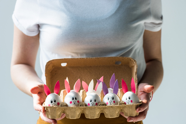 vista parcial de mujer sosteniendo bandeja de huevo con huevos de gallina con caras divertidas de conejito y orejas de papel aisladas en blanco aisladas en gris
 - Foto, Imagen