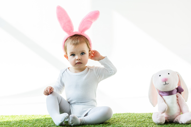 白い上に孤立したおもちゃのウサギの近くに座っているバニーの耳のカチューシャのかわいい子供 - 写真・画像