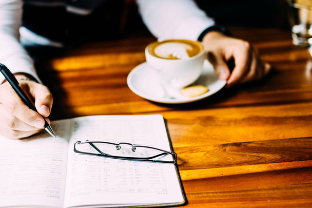 La mano de un hombre sentado en una mesa con una taza de café aromático, un teléfono y un cuaderno. Almuerzo de negocios
 - Foto, imagen